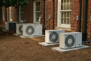 A row of five white outdoor air conditioning condenser units installed on concrete pads outside a brick apartment building.