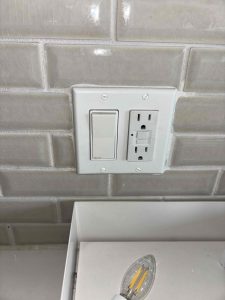 A close-up of a white double-gang wall plate mounted on a glossy grey subway tile kitchen backsplash, featuring a modern rocker light switch alongside a GFCI electrical outlet.