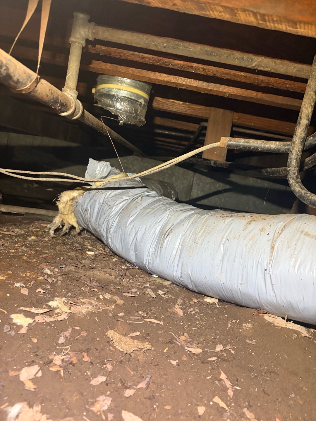 View underneath a house in a crawlspace showing old cloth electrical wiring hanging near pipes and flexible ductwork lying on the dirt ground.