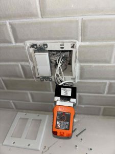 An exposed electrical wall box on a kitchen backsplash during troubleshooting, showing a Klein Tools RT210 receptacle tester plugged into a disassembled GFCI outlet with visible wiring.